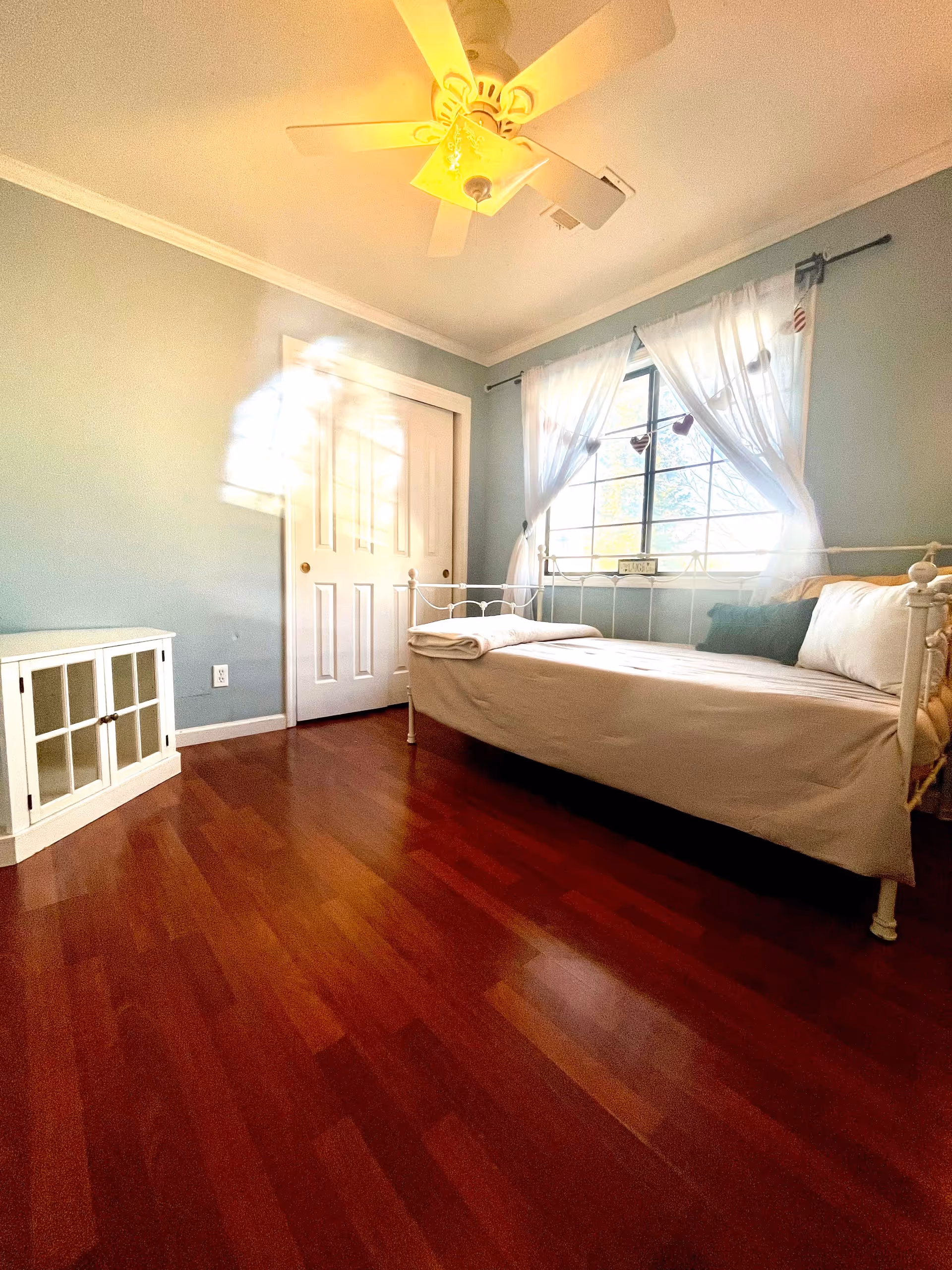 A bright bedroom with light blue walls, a white metal daybed with white and green pillows, a folded blanket on the bed, a window with sheer white curtains letting in natural light, a white cabinet with glass doors, a ceiling fan with a light, and a closed white closet door.