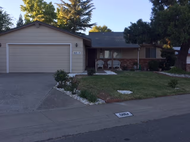 Front exterior view of a single-story residential building with a two-car garage, a small front porch with two chairs, a lawn with some shrubs and trees, and the house number 6813 displayed on the garage and curb.