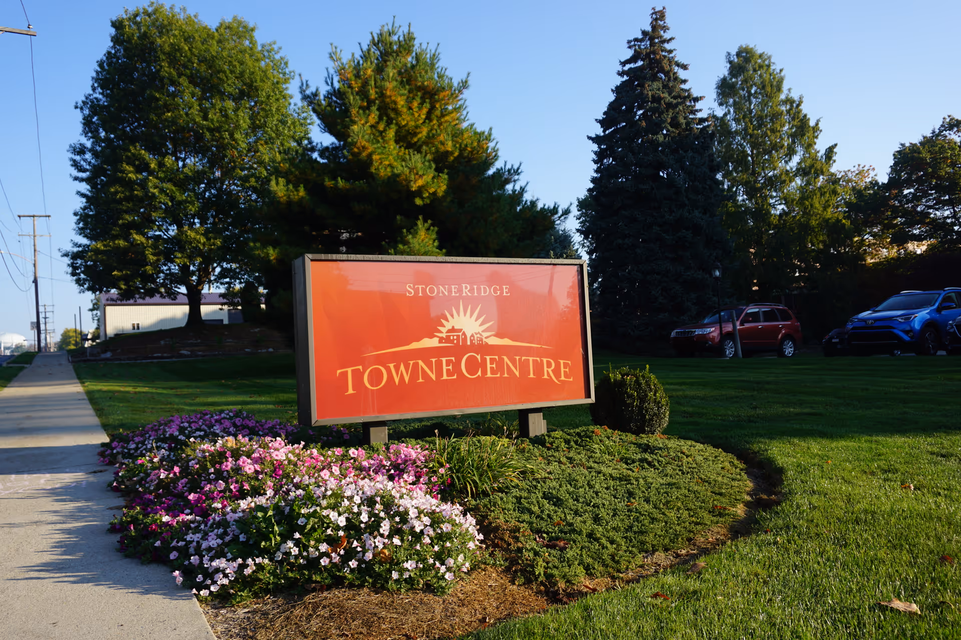 Outdoor view of a landscaped area with a red sign that reads 'StoneRidge Towne Centre' surrounded by green grass, bushes, and colorful flowers, with trees and parked cars in the background under a clear blue sky.