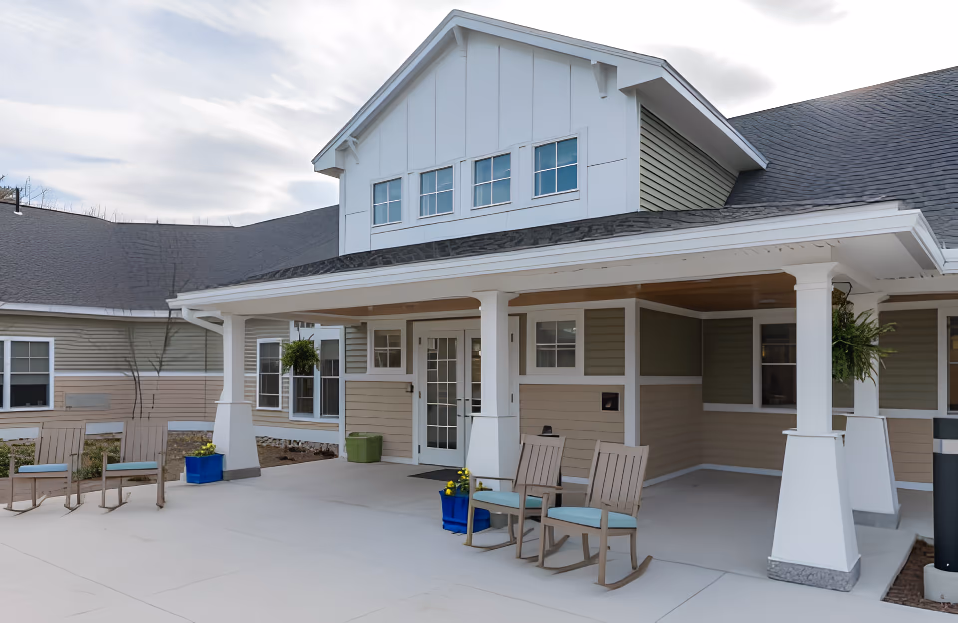 Exterior view of a senior living facility entrance with a covered porch, two rocking chairs with cushions, potted plants, and multiple windows under a cloudy sky.