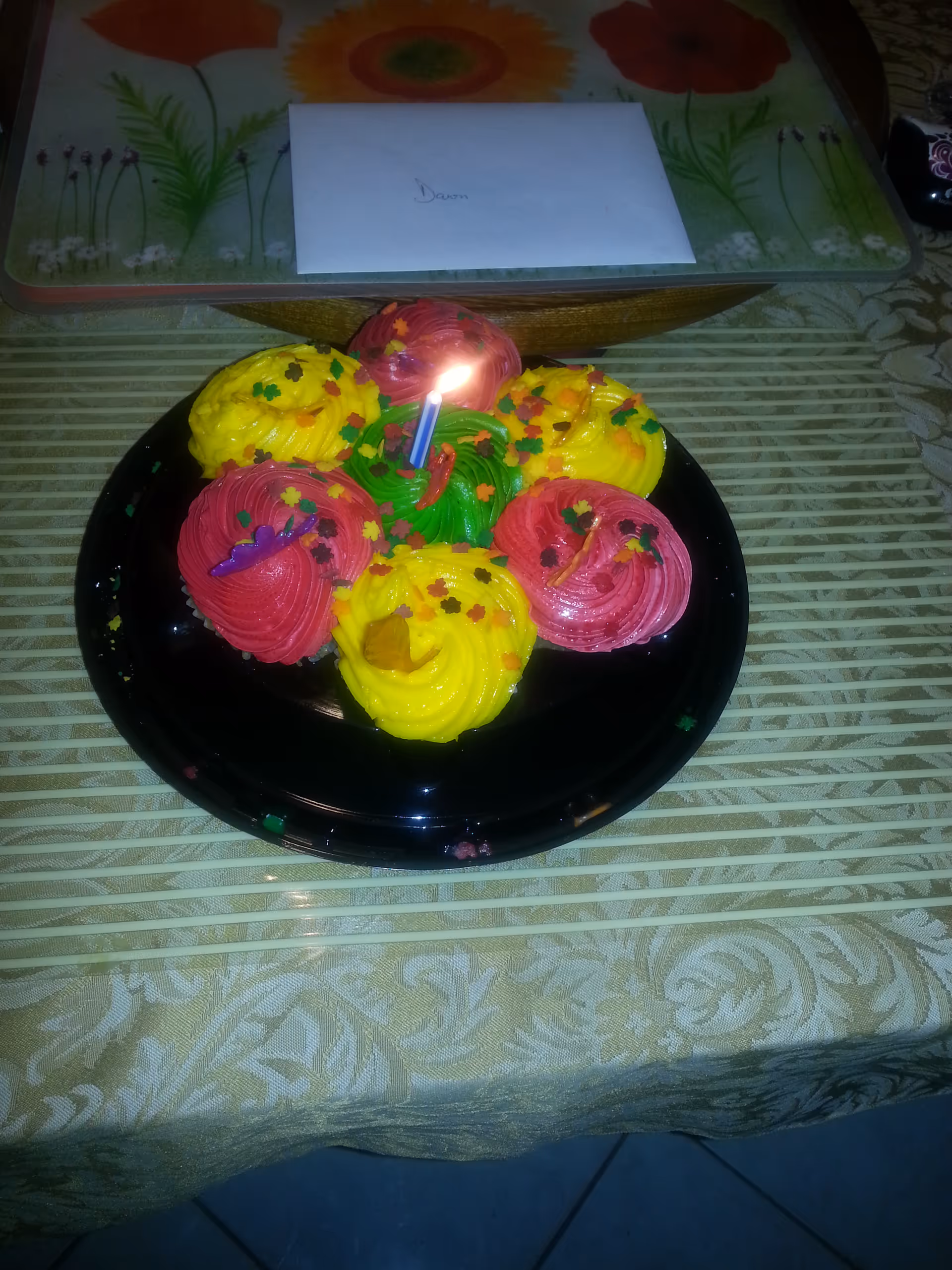 A plate with six colorful cupcakes decorated with yellow, red, and green frosting and small leaf-shaped sprinkles. One cupcake has a lit candle. The plate is on a patterned tablecloth with a floral design in the background and an envelope placed on a glass surface behind the cupcakes.