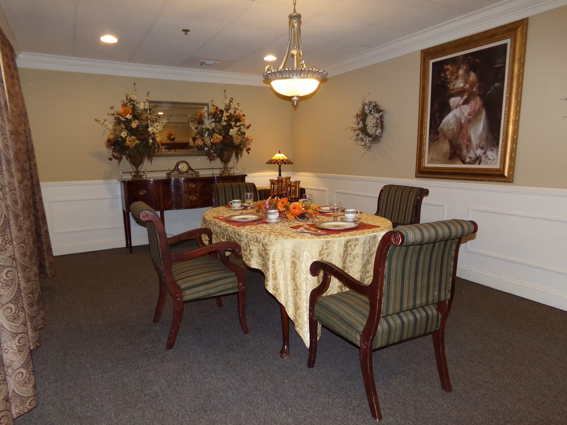 A formal dining room with a round table set for four, upholstered chairs, a sideboard with floral arrangements, and framed artwork on the walls.