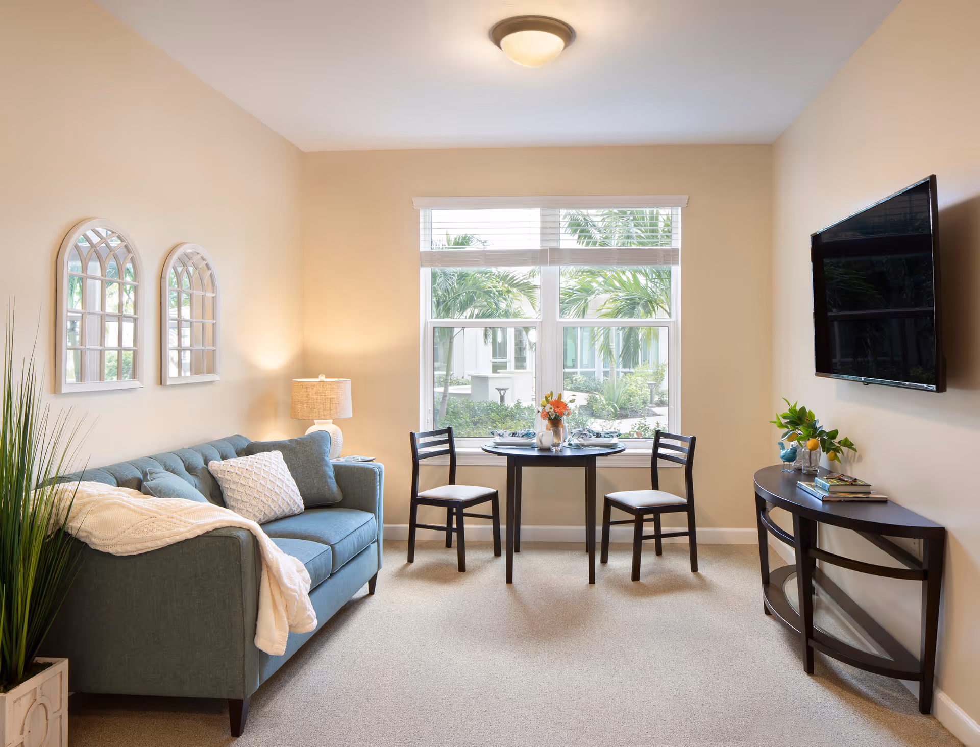 A cozy living room with a blue sofa adorned with pillows and a white throw blanket. Two decorative mirrors hang on the wall above the sofa. A small round dining table with two chairs is placed near a large window showing greenery outside. A flat-screen TV is mounted on the wall opposite the sofa, above a dark wooden console table with books and a plant. The room is softly lit by a ceiling light and a table lamp beside the sofa.