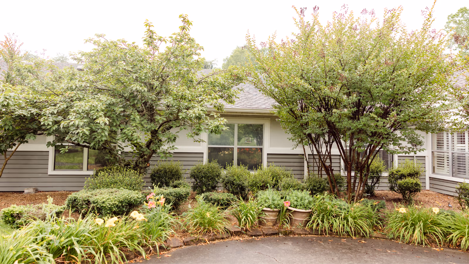 A landscaped garden area in front of a single-story building with gray siding and white trim. The garden features various green shrubs, flowering plants, and two medium-sized trees. The building has multiple windows with white frames.