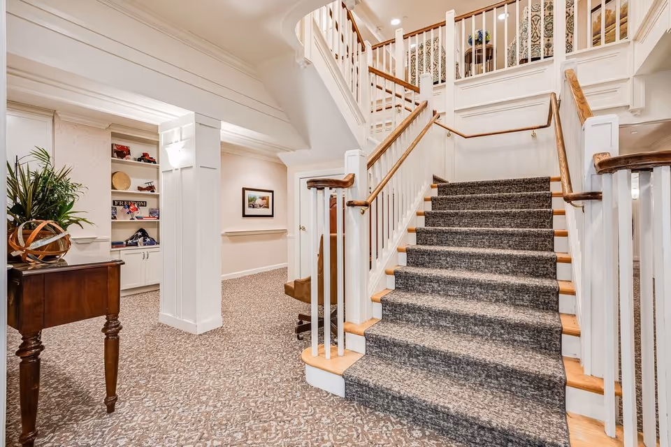 Interior view of a senior living facility showing a carpeted staircase with wooden handrails leading to an upper floor. To the left, there is a small wooden table with a decorative plant and a built-in shelving unit with various decorative items. The walls are light-colored with a framed picture hanging, and the floor is carpeted with a patterned design.
