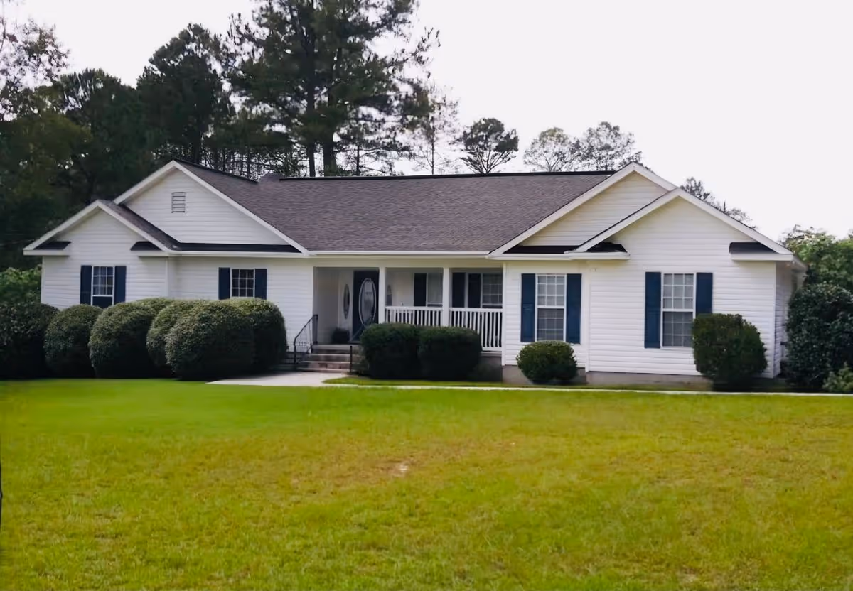 Single-story white house with a front porch, blue shutters, shrubs, and a large lawn.