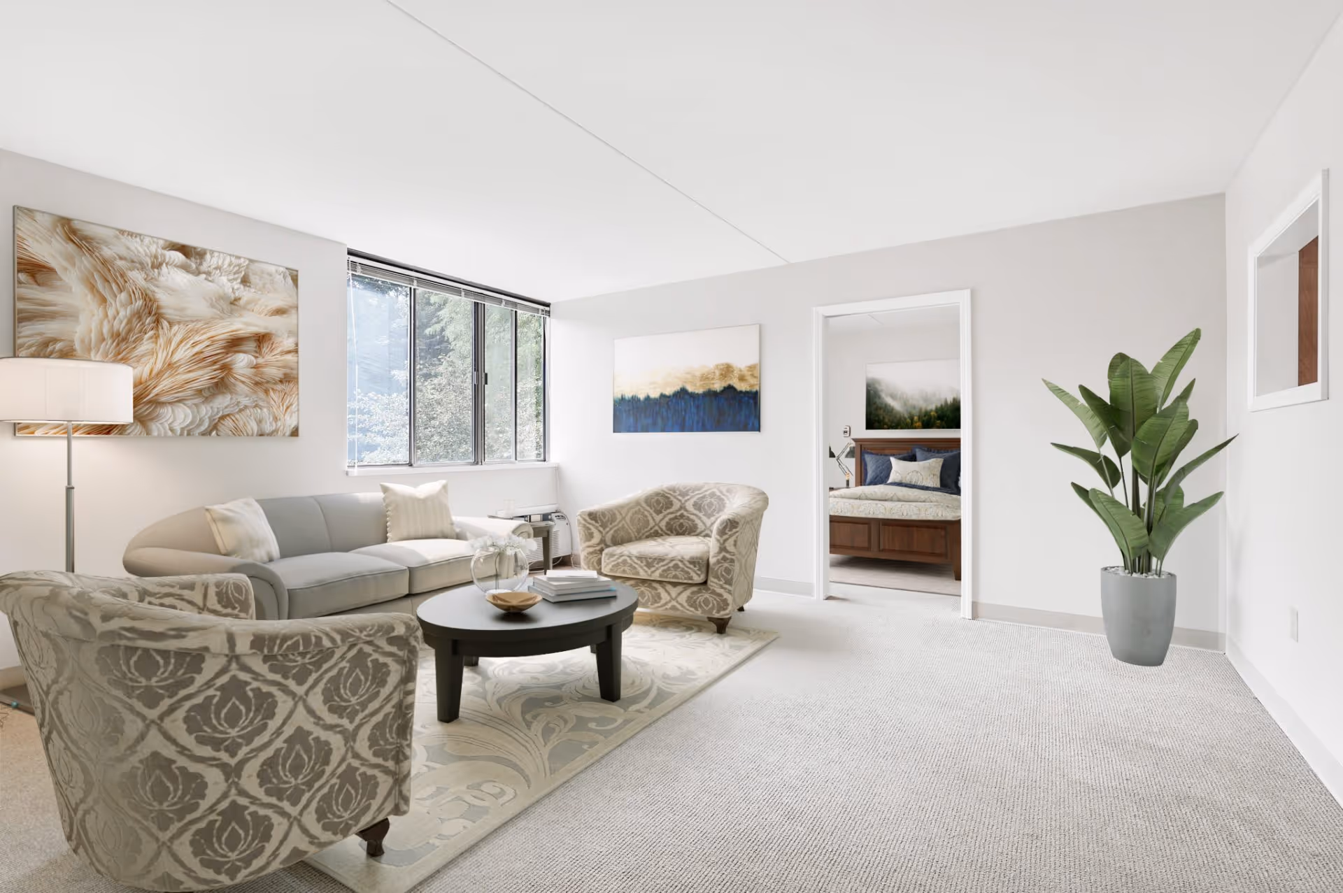 A bright and spacious living room with a large window letting in natural light. The room features a light gray sofa, two patterned armchairs, a round black coffee table with books and a decorative bowl, a floor lamp, and two abstract paintings on the walls. A doorway leads to a bedroom with a wooden bed frame and blue bedding. There is also a large green potted plant in the corner.