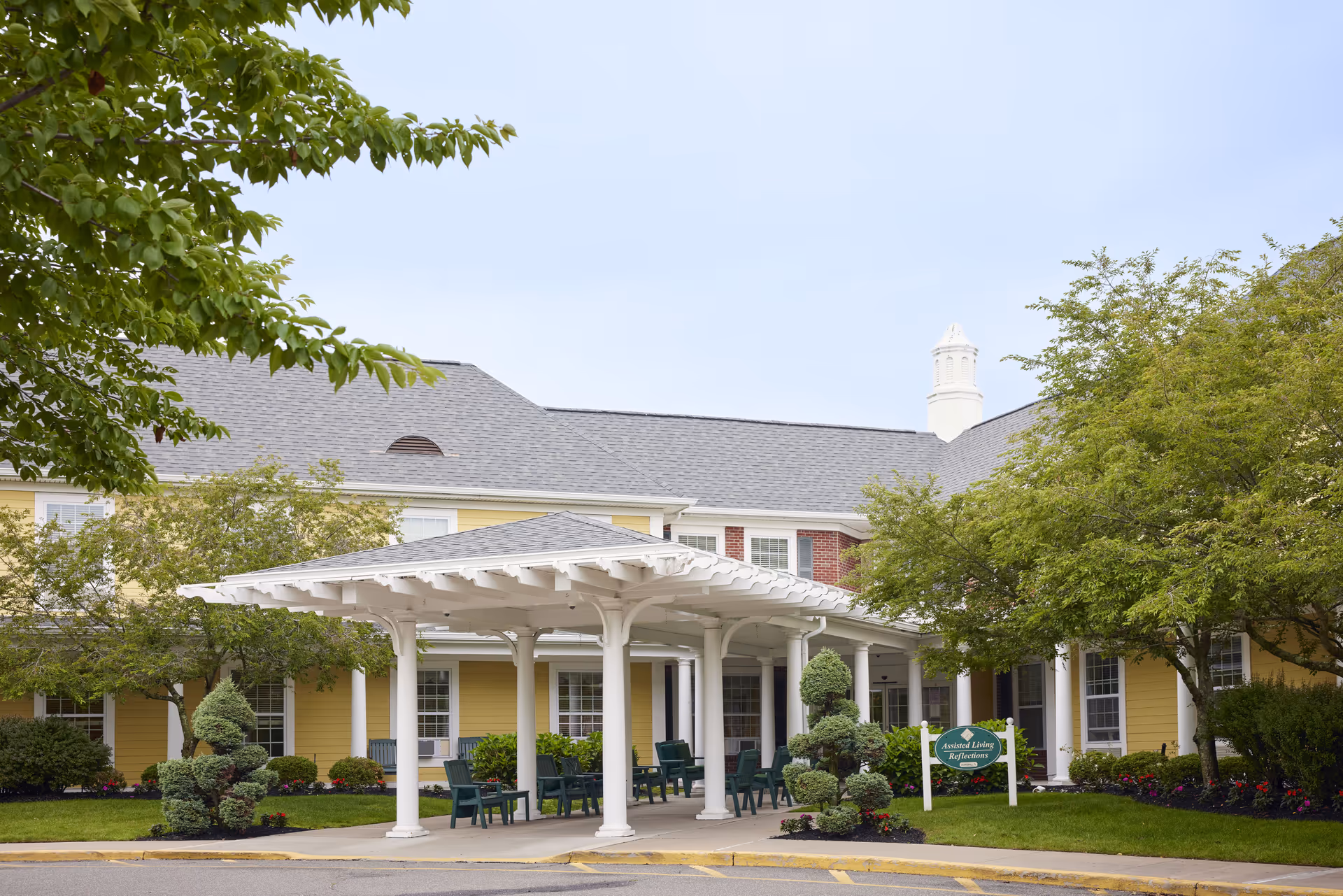 Front entrance of a yellow senior living building featuring a white porte-cochere, green chairs, and landscaped trees.