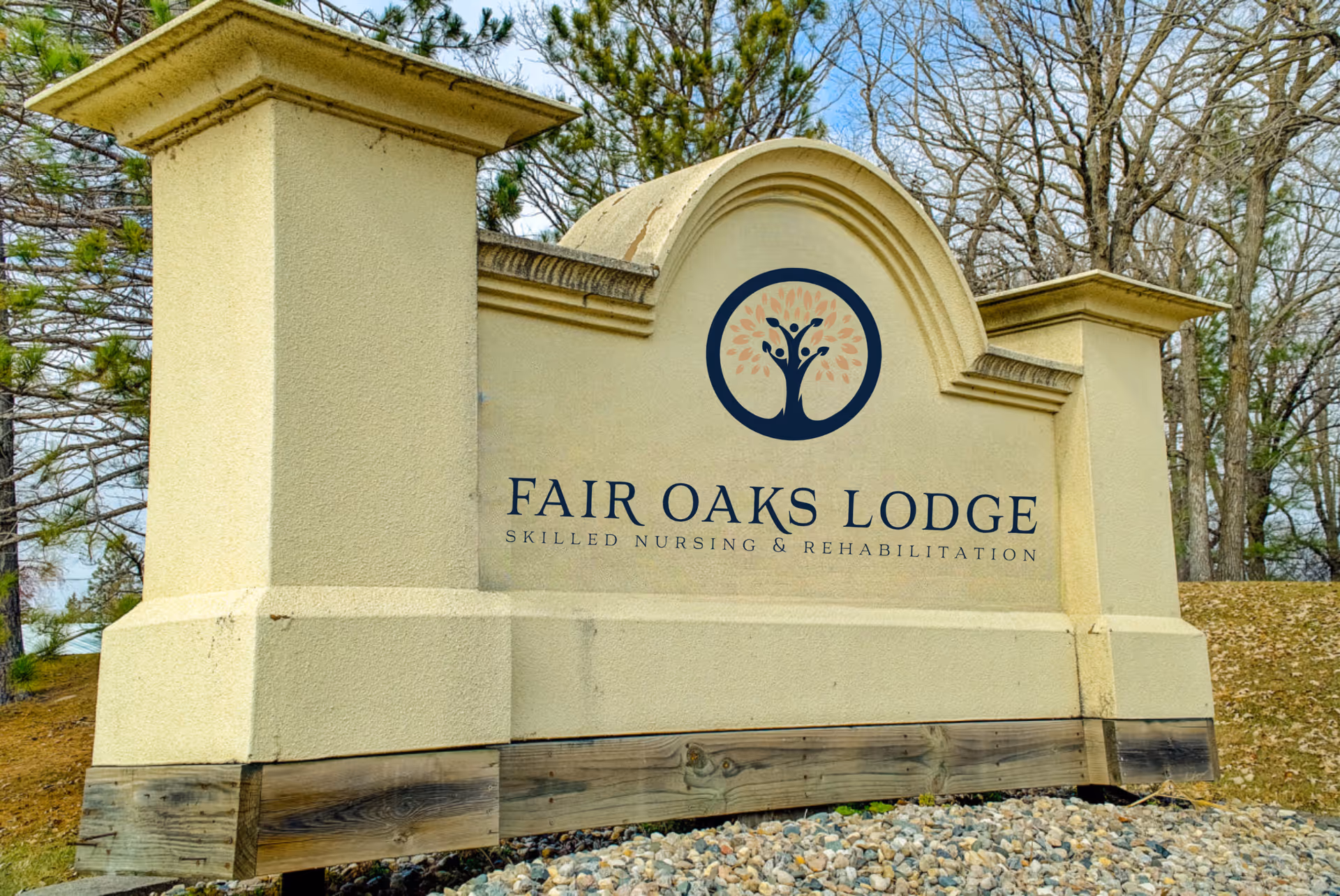 A large beige stone sign for Fair Oaks Lodge Skilled Nursing & Rehabilitation with a tree logo, set outdoors with trees and grass in the background.