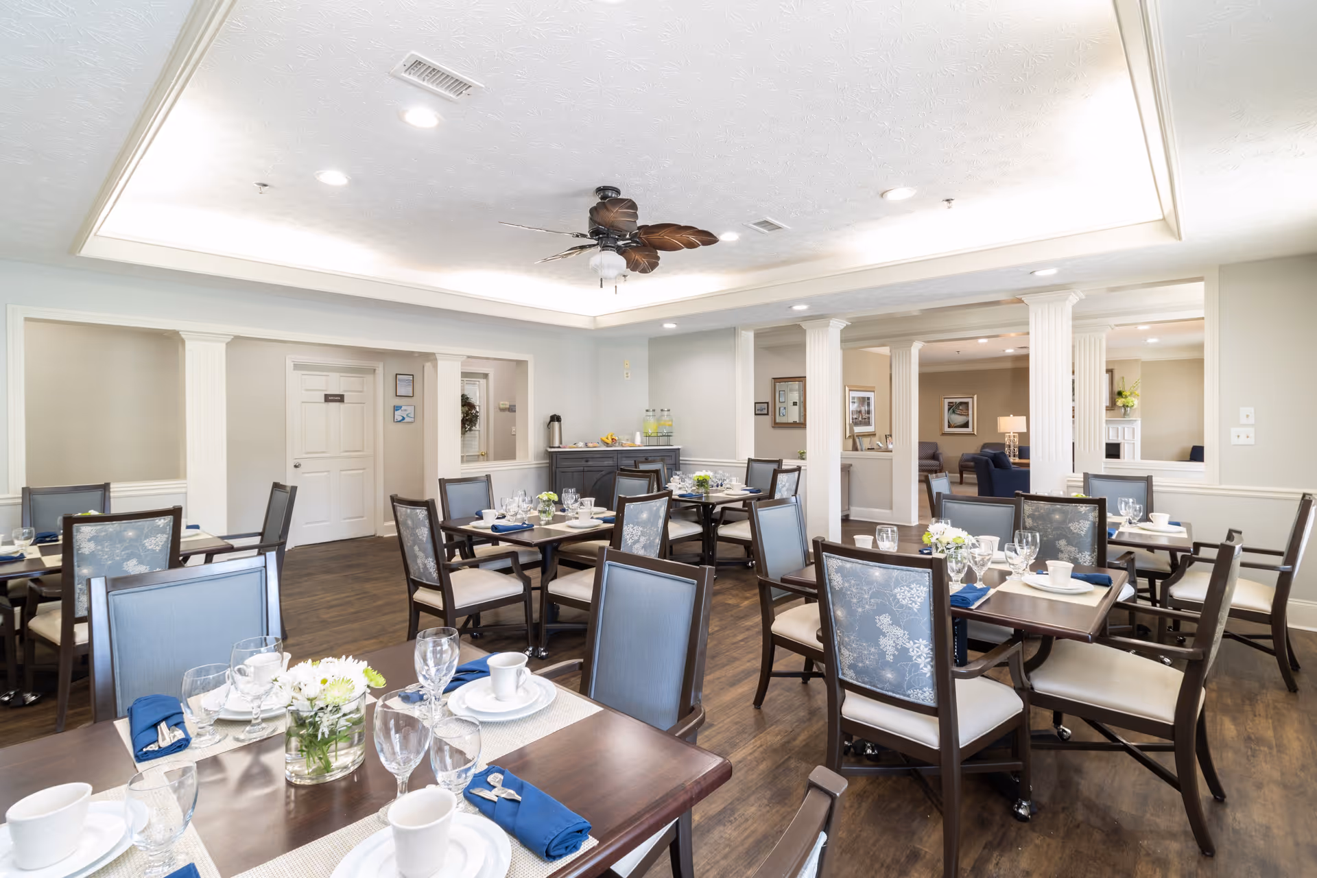A bright dining room in a senior living facility with multiple tables set with white plates, cups, glassware, and blue napkins. The room features wooden floors, light-colored walls, decorative columns, and a ceiling fan with leaf-shaped blades. There is a sideboard with water bottles and a coffee dispenser in the background, and a lounge area is visible through an open wall space.