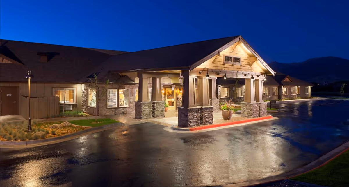 Exterior view of a senior living facility entrance at night with warm lighting illuminating the covered entryway and surrounding building. The driveway is wet, reflecting the lights, and there are mountains visible in the background under a clear dark blue sky.