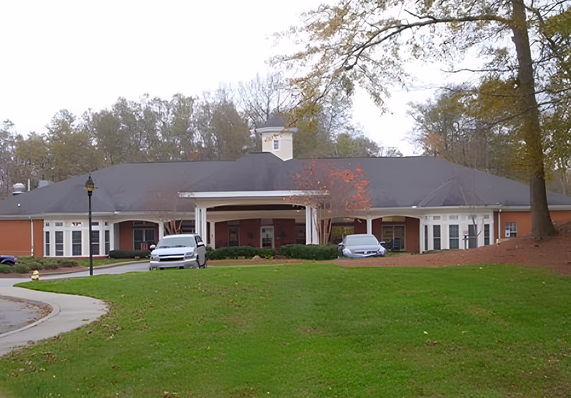 Front view of a single-story brick senior living building with a covered drive-up entrance, parked cars, and a grassy lawn.