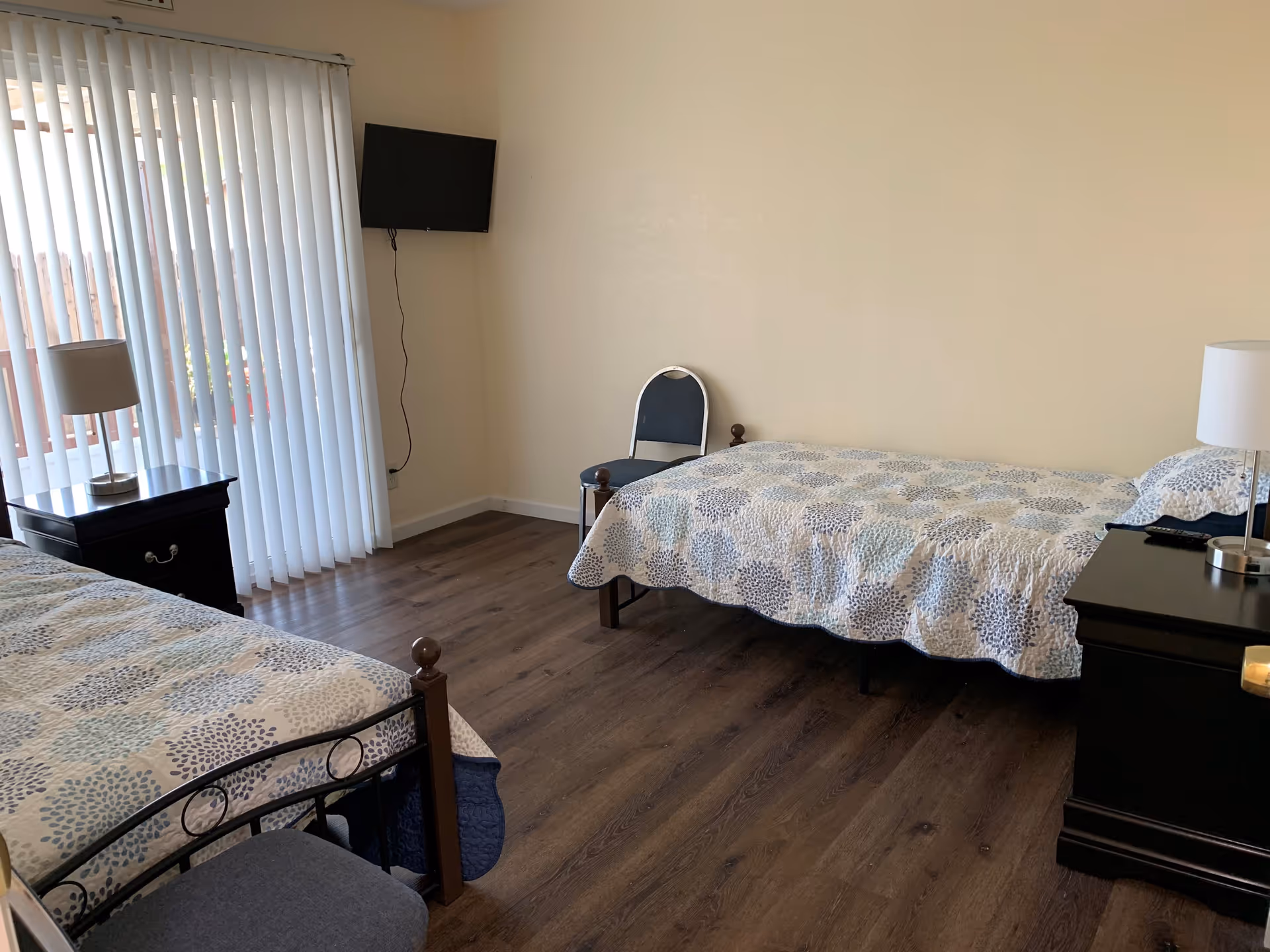 A bedroom with two single beds covered with patterned bedspreads, a black nightstand with a lamp on each side, a chair against the wall, a wall-mounted TV, and vertical blinds covering a sliding glass door.