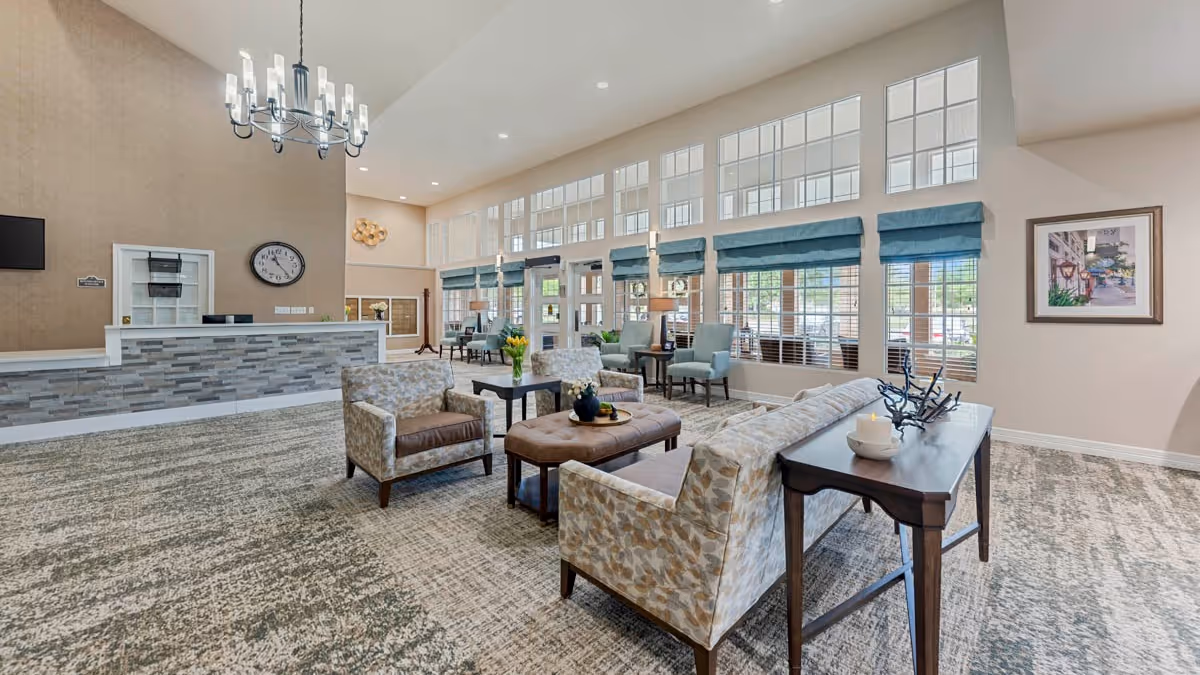 Spacious senior living facility lobby with upholstered seating, a central coffee table, reception desk, and large windows with high ceilings.