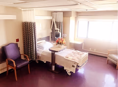 A bright and clean hospital-style room with a single bed, a bedside table with a flower arrangement, a purple chair, and a large window letting in natural light. The room has light-colored walls and purple flooring.