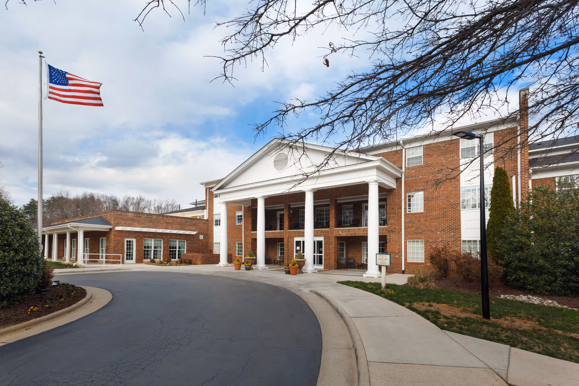Exterior view of a senior living facility named Abbotswood at Irving Park, featuring a brick building with white columns at the entrance, an American flag on a flagpole, and a curved driveway leading to the entrance. Trees and shrubs surround the building under a partly cloudy sky.