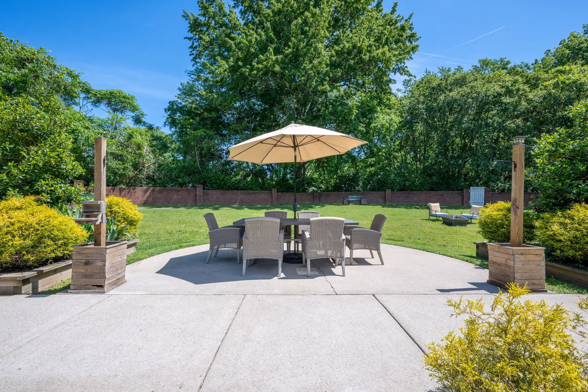 Outdoor patio area with a round table surrounded by six wicker chairs under a large beige umbrella. The patio is surrounded by green grass, bushes, and trees with a clear blue sky overhead.