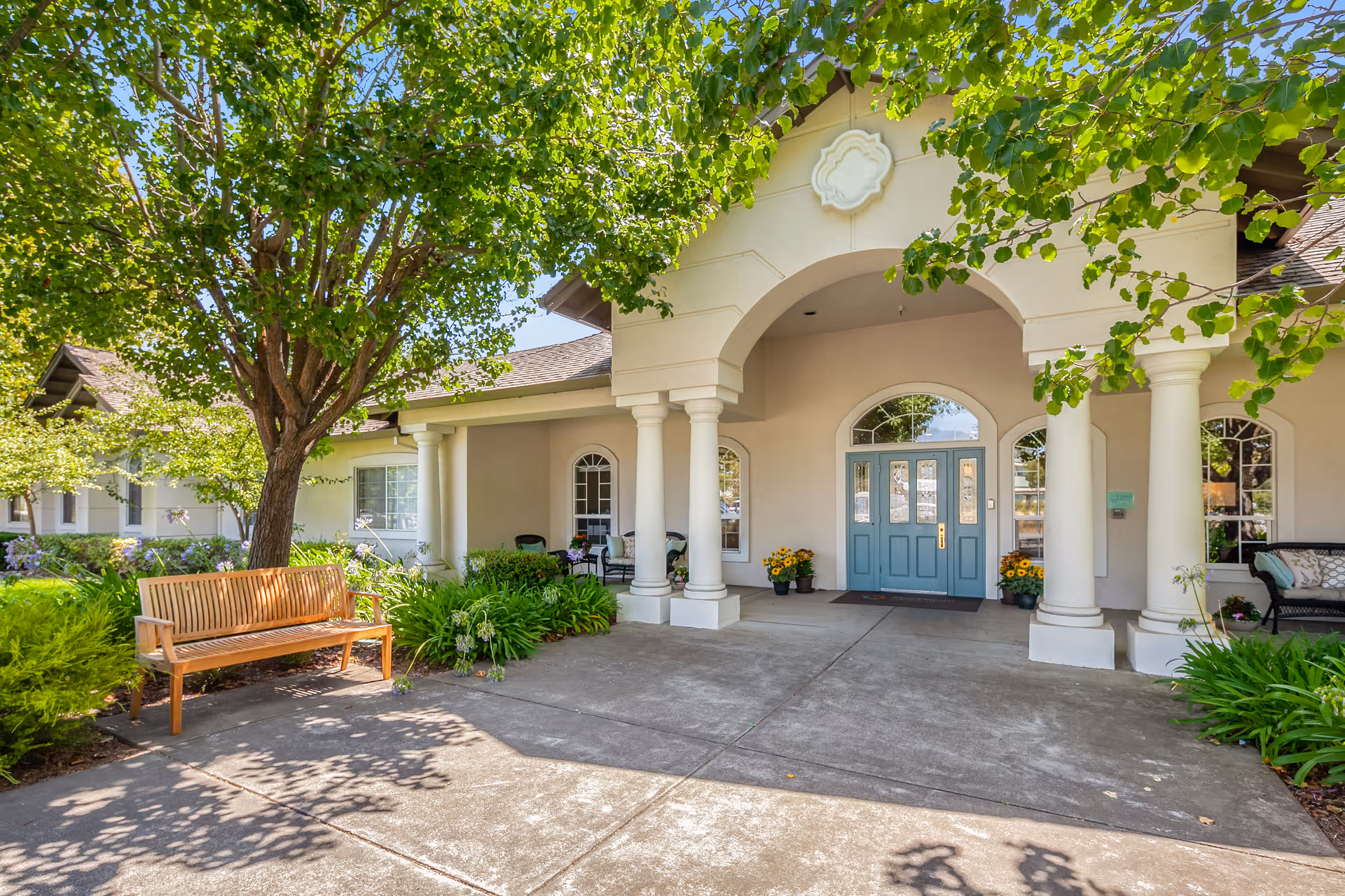 Entrance of MuirWoods Memory Care facility featuring a covered porch with white columns, a blue double door, potted plants with yellow flowers, a wooden bench under a leafy tree, and additional seating on the porch surrounded by greenery.