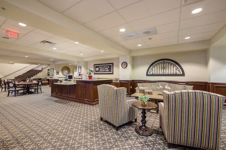 Lobby-style common area with seating, a reception desk, dining tables, and a staircase in a senior living facility.