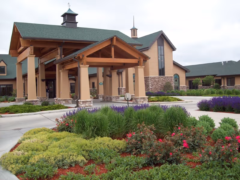 Entrance of a senior living facility with a covered driveway supported by large wooden beams and stone pillars. The building has beige siding with stone accents and green roofing. In front, there is a landscaped garden with various green shrubs, purple flowers, and red roses.