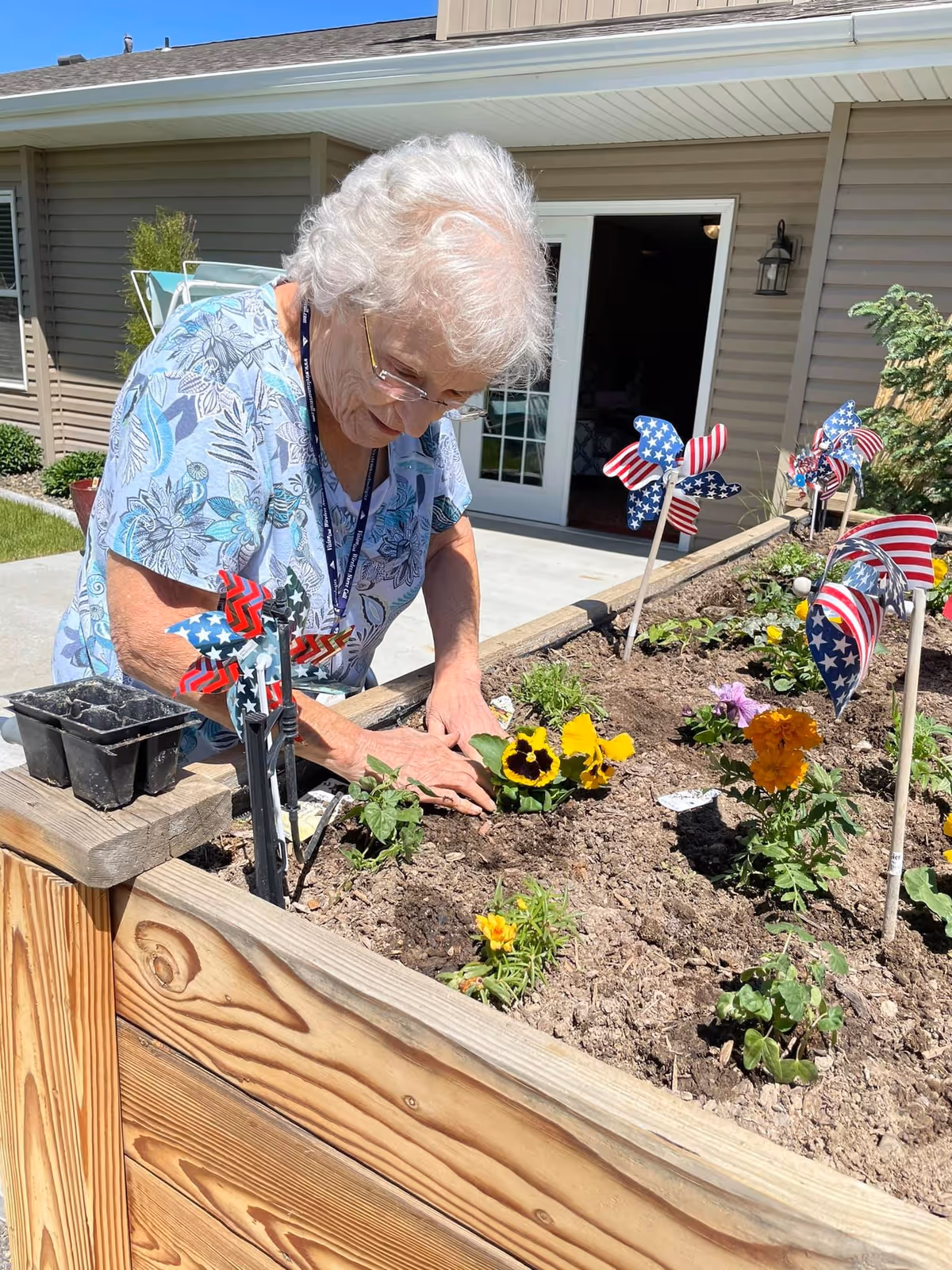 An elderly woman with white hair and glasses is gardening in a raised wooden flower bed outside a building. She is planting yellow and orange flowers in the soil. The flower bed is decorated with several pinwheels featuring American flag designs. The building behind her has beige siding, a white door, and a window.