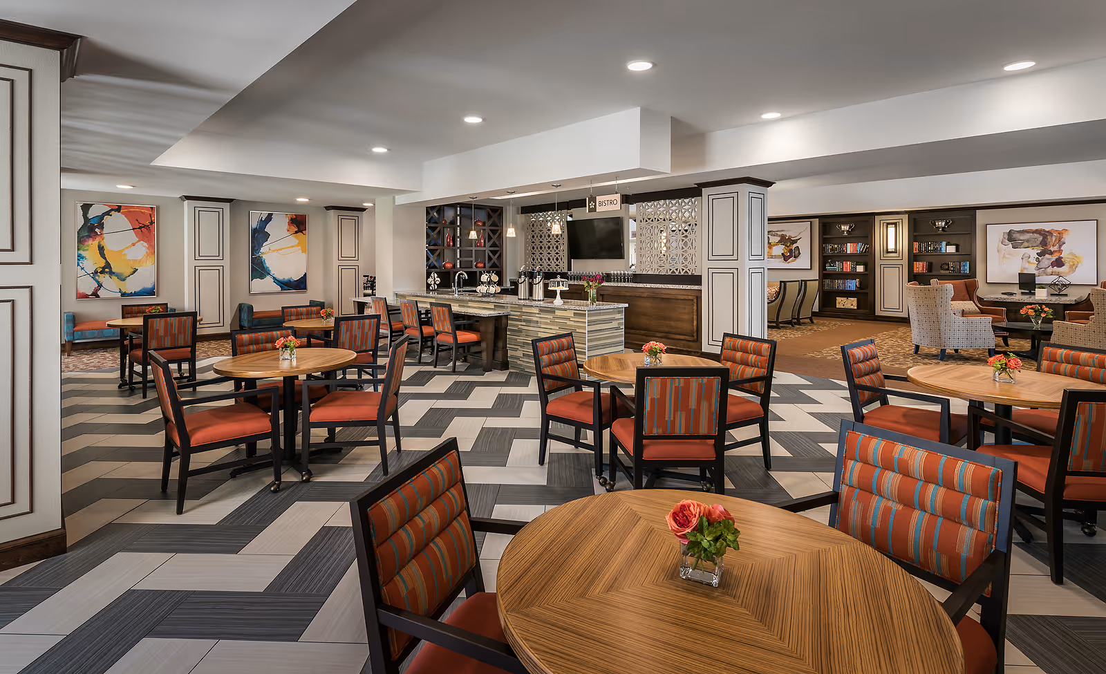 Open communal dining and bistro area with round wooden tables, upholstered chairs, and a service counter in a well-lit interior.