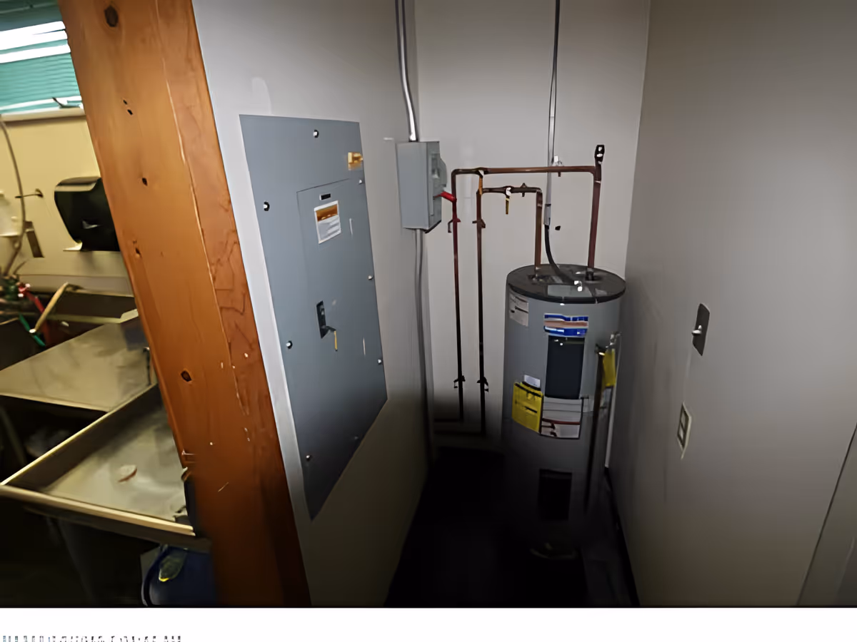 Utility room with a water heater, electrical panel, and some exposed pipes. Part of a stainless steel sink and a paper towel dispenser are visible in an adjacent room.