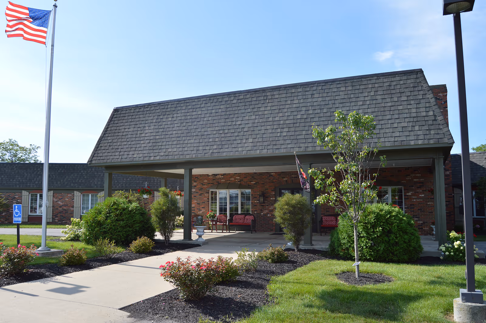 Front exterior view of Van Wert Manor, a single-story brick building with a covered entrance, landscaped bushes and flowers, an American flag on a flagpole, and a sidewalk leading to the entrance.