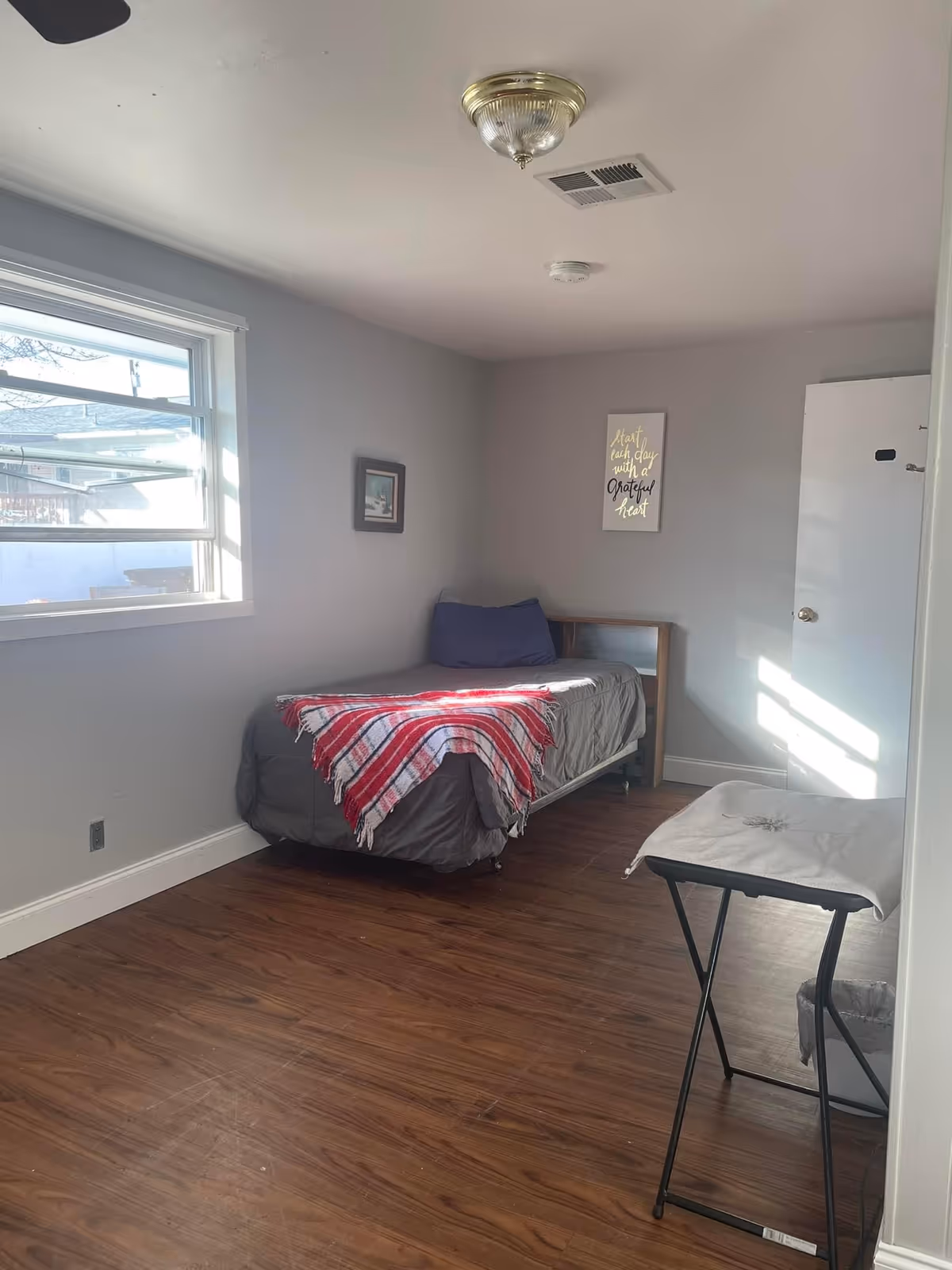 A small bedroom with a single bed against the wall, covered with a gray bedspread and a red and white striped blanket. There is a blue pillow on the bed. The room has wooden flooring, a window letting in natural light, a small framed picture on the wall, and a wall decoration with the text 'Start each day with a grateful heart'. A small folding table with a cloth cover is positioned near the door.