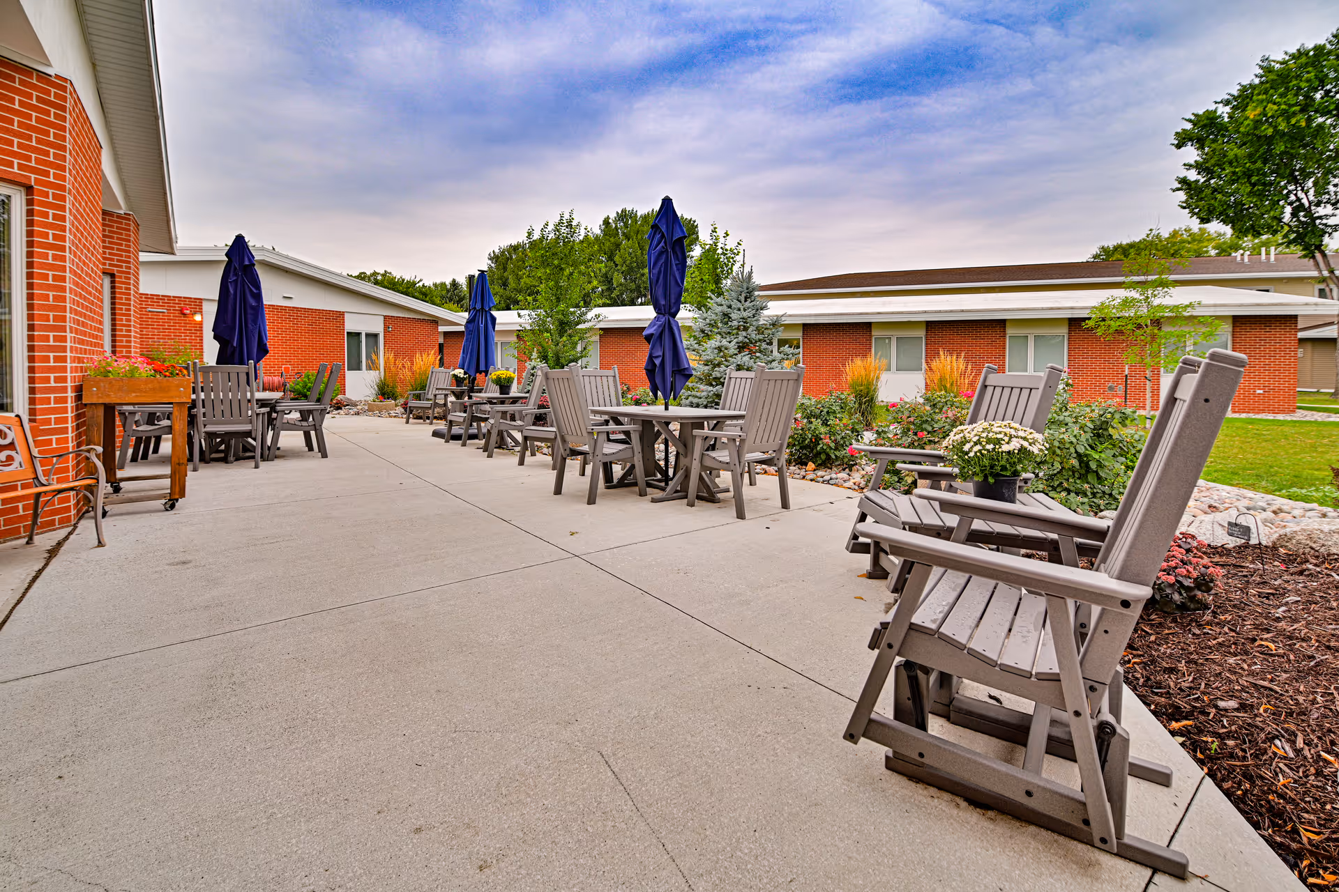 Outdoor patio area at Valley Senior Living on Cherry - Tufte Manor with multiple wooden chairs and tables, some with closed blue umbrellas. The patio is surrounded by landscaped garden beds with flowers and shrubs, and red brick buildings are visible in the background under a partly cloudy sky.