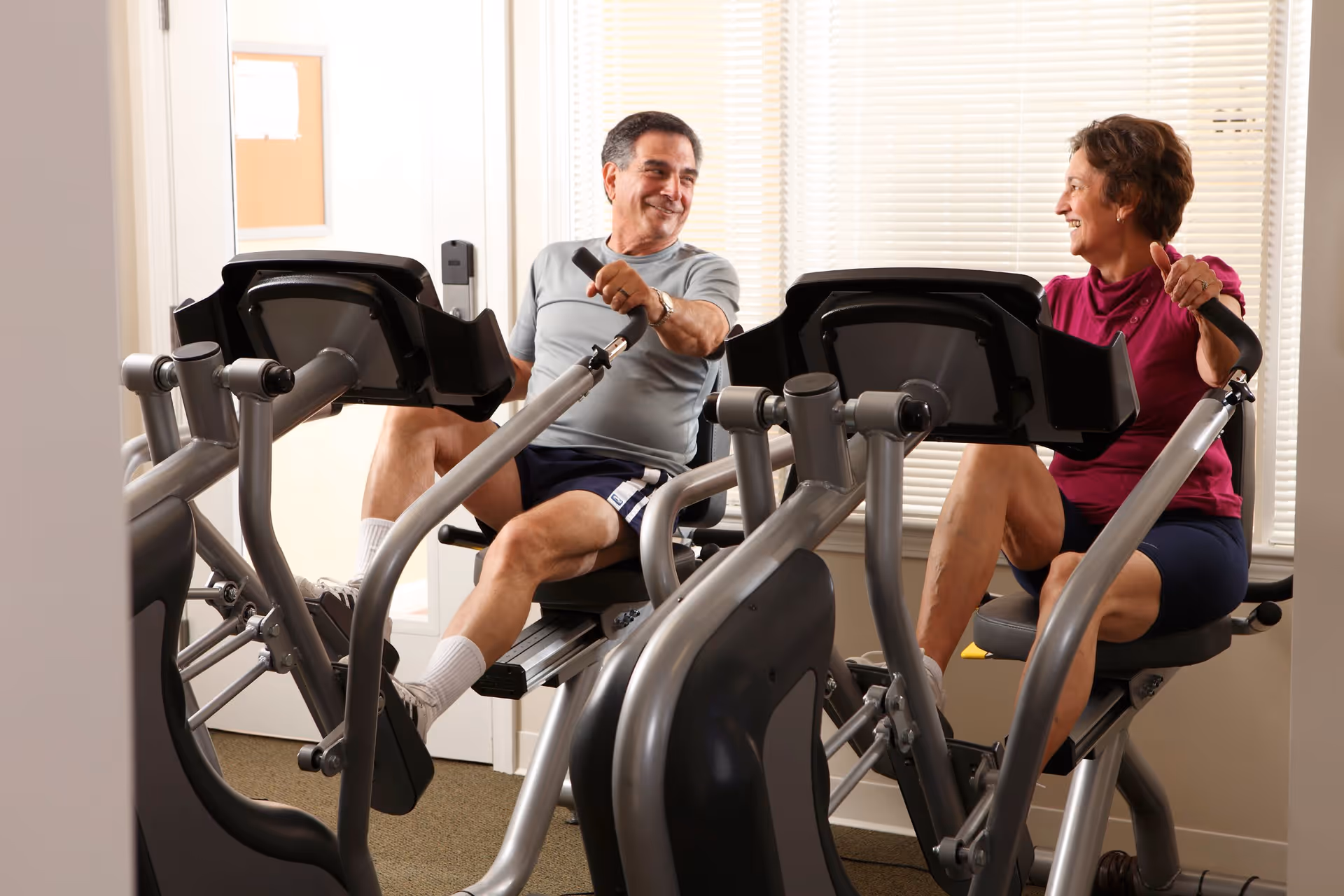 An elderly man and woman exercising on seated rowing machines in a fitness room with large windows and blinds, smiling and looking at each other.