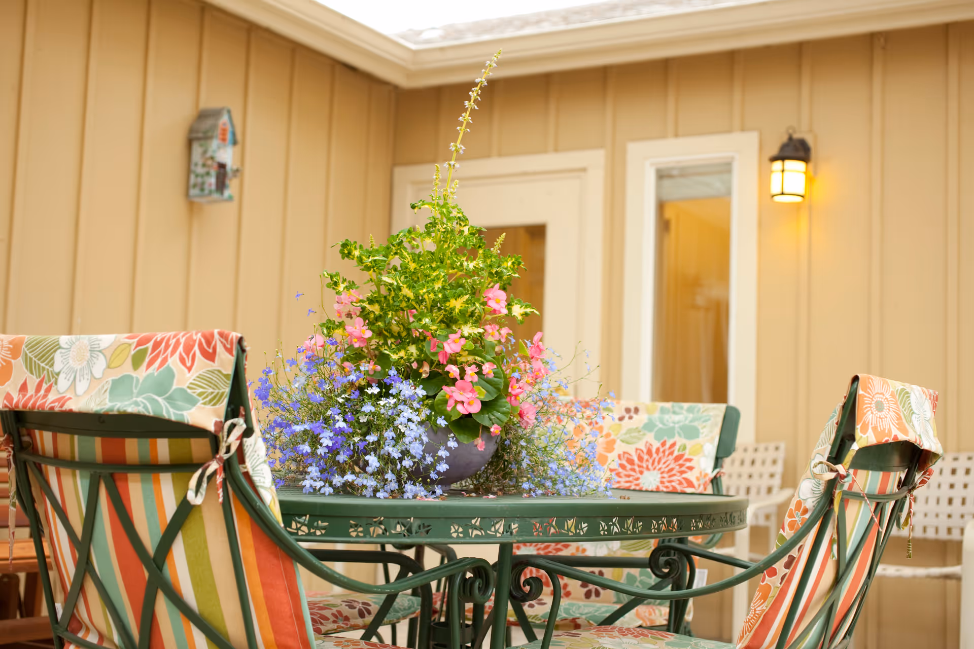 Outdoor patio area with a green metal table and chairs featuring colorful floral and striped cushions. A large flower arrangement with pink, purple, and green plants sits on the table. The background shows a beige building wall with a door, window, a lit wall lantern, and a decorative birdhouse.