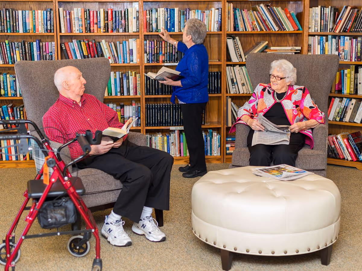 Three elderly residents reading in a library lounge with bookshelves, armchairs, an ottoman, and a walker.