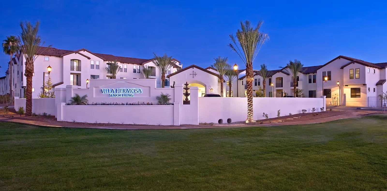 Exterior view of Villa Hermosa senior living facility at dusk, featuring white buildings with red tile roofs, palm trees, illuminated street lamps, and a well-maintained green lawn in the foreground.