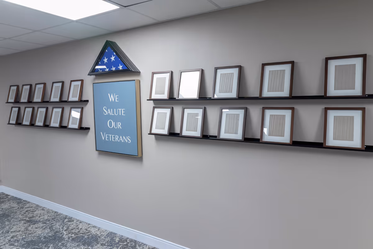 A hallway wall display featuring a folded American flag in a triangular case above a blue sign that reads 'We Salute Our Veterans,' surrounded by multiple empty picture frames on two black shelves.