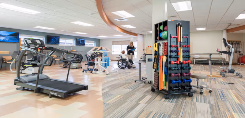 A spacious physical therapy or rehabilitation room with exercise equipment including a treadmill, weights, resistance bands, parallel bars, and therapy tables. A therapist is assisting a person in a wheelchair near the back of the room. The room has bright lighting and a mix of patterned and solid flooring.