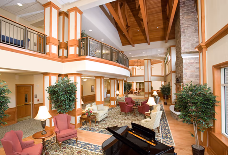 Spacious and well-lit senior living facility common area with high wooden ceilings, a stone fireplace, multiple seating arrangements including armchairs and sofas, potted plants, table lamps, and a black grand piano in the foreground.