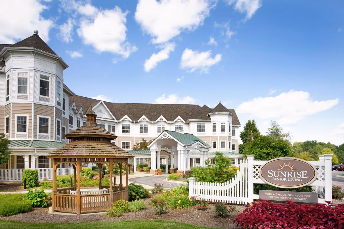 Exterior view of Sunrise of Naperville North senior living facility featuring a large multi-story building with white and beige siding, a covered entrance, a wooden gazebo in the landscaped garden area, and a white picket fence with a sign that reads 'Sunrise Senior Living - With a Special Neighborhood for the Memory Impaired'. The sky is blue with scattered clouds.