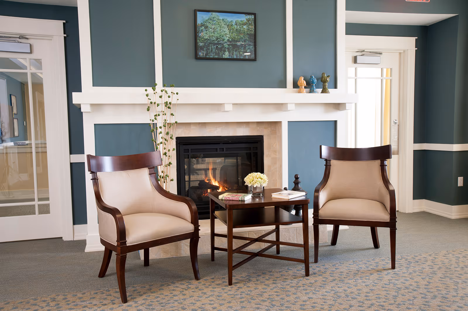 A cozy seating area with two beige upholstered wooden armchairs facing a small wooden coffee table. Behind the table is a lit fireplace with a white mantel, decorated with small figurines and a framed landscape painting above it. The walls are painted teal with white trim, and there is a door with glass panels on the left side.