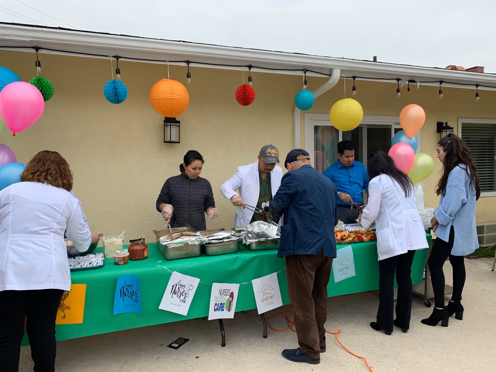 Staff and residents serving food at a decorated outdoor buffet table against a beige building with colorful balloons and paper lanterns.
