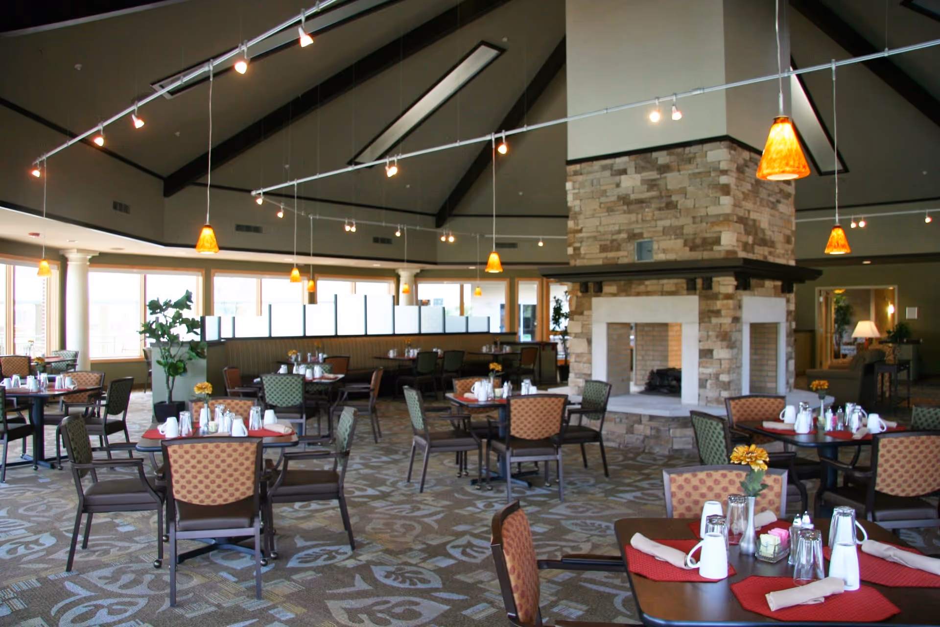 Spacious dining room with multiple tables and chairs arranged neatly, each table set with white mugs, glasses, napkins, and small flower centerpieces. The room features a large stone fireplace in the center, high vaulted ceilings with exposed beams, and pendant lights hanging from the ceiling. Large windows allow natural light to fill the space.