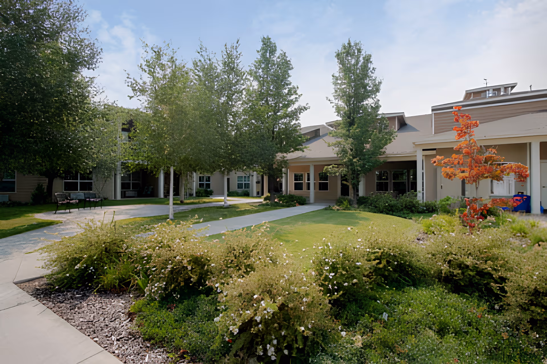 Exterior view of Wenatchee Senior Living by Cogir showing a landscaped garden with bushes, trees, and a walkway leading to the building entrance under a partly cloudy sky.