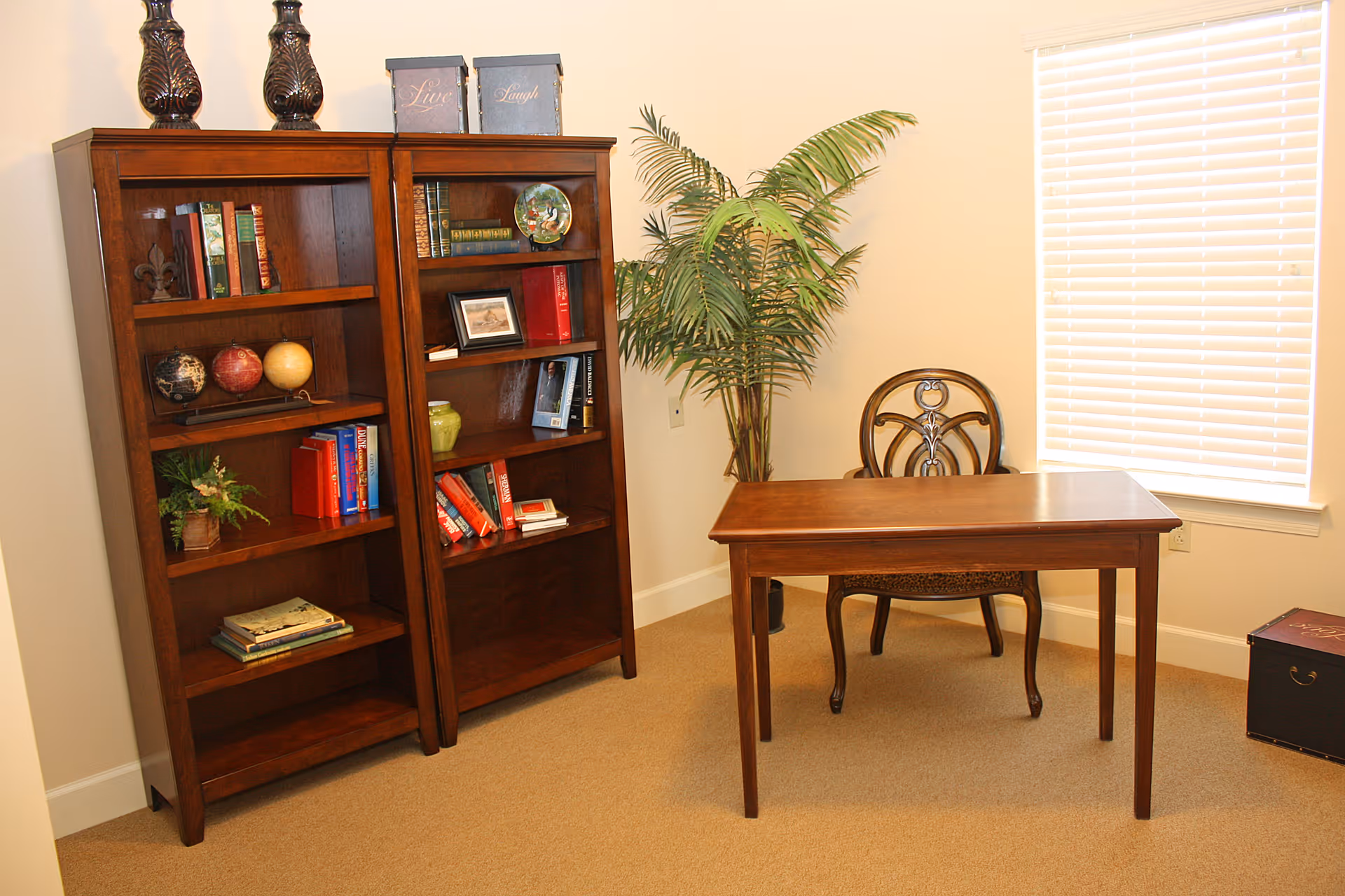 A small office or study area with two wooden bookshelves filled with books and decorative items, a wooden desk with a matching chair, a potted plant in the corner, and a window with closed blinds.