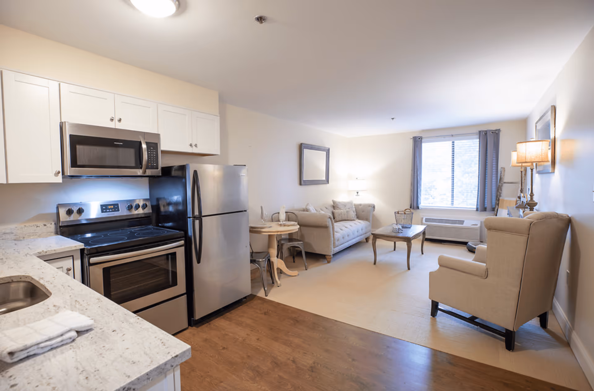 A bright and clean living space featuring a modern kitchen with stainless steel appliances including a microwave, stove, and refrigerator. Adjacent to the kitchen is a cozy living room area with a beige sofa, armchair, coffee table, and a window with curtains letting in natural light. The room has light-colored walls and wooden flooring near the kitchen area transitioning to carpet in the living area.