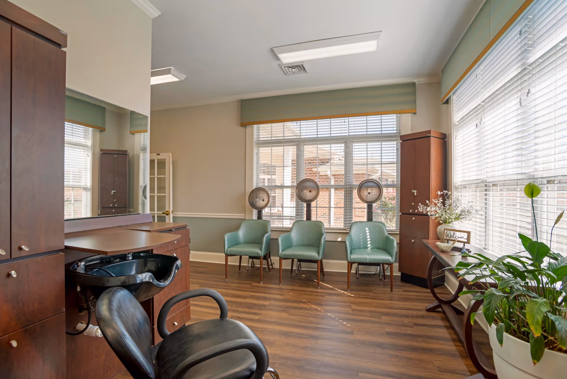 A bright and clean hair salon area in a senior living facility with three green salon chairs facing hair dryers near large windows with blinds. There is a black salon chair in front of a wooden counter with a sink, and a wooden cabinet on the left. A small table with a plant and a welcome sign is on the right side near another window.