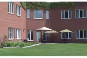Brick senior living building courtyard with a green lawn, two patio tables with umbrellas, and ground-floor windows.