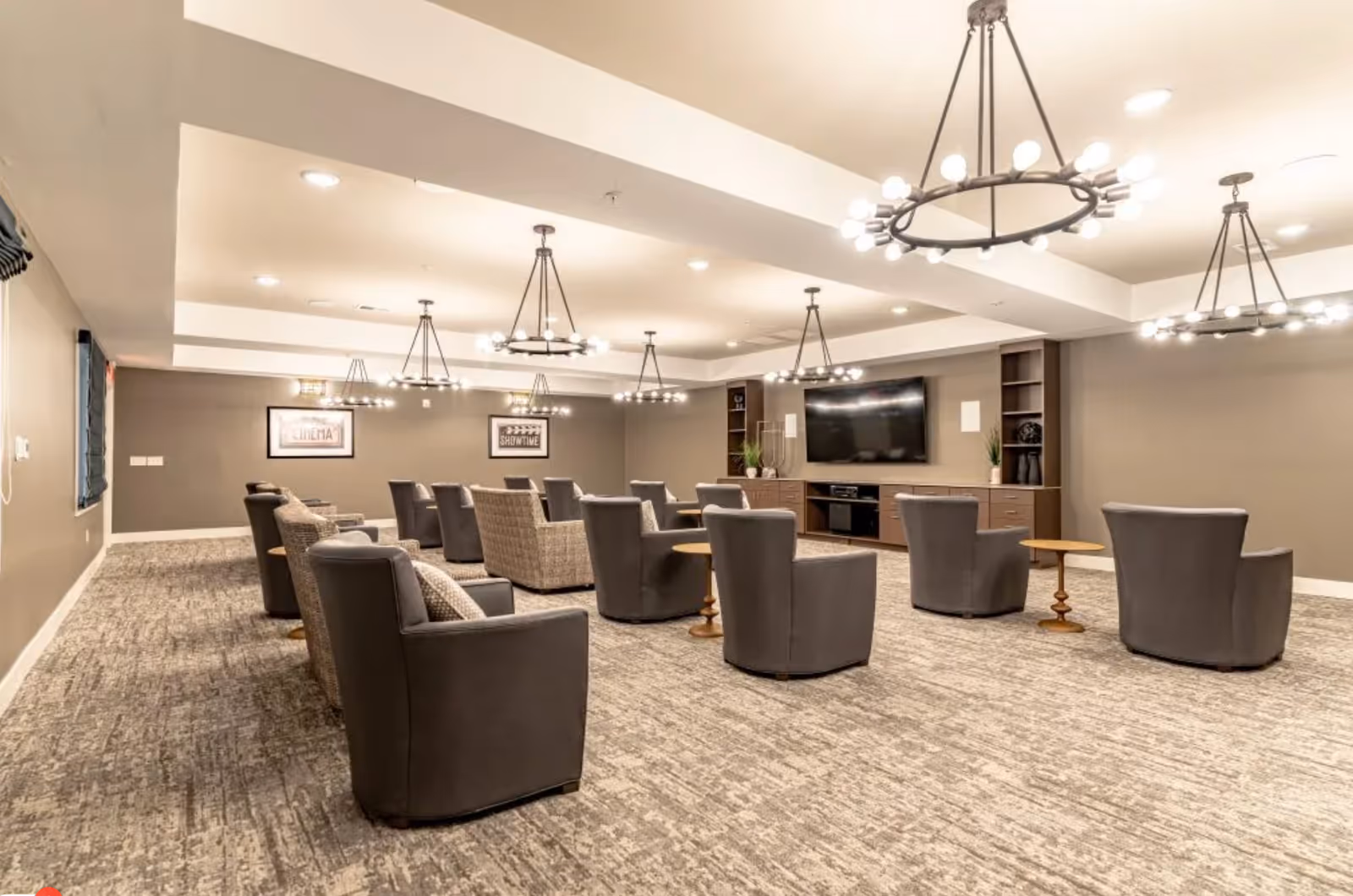A spacious, well-lit common area with multiple comfortable armchairs arranged in rows facing a large flat-screen TV mounted on a wall with built-in shelving. The room has modern chandeliers, neutral-colored walls, and carpeted flooring, creating a cozy and inviting atmosphere.