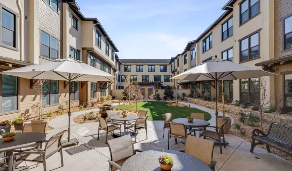 Outdoor courtyard with patio tables, umbrellas, and a central lawn surrounded by a three-story residential building.
