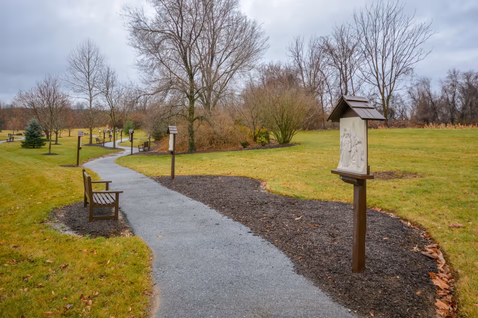 A winding paved pathway through a grassy outdoor area with several wooden benches and small wooden stands displaying plaques or artwork. Leafless trees and bushes surround the path under a cloudy sky.