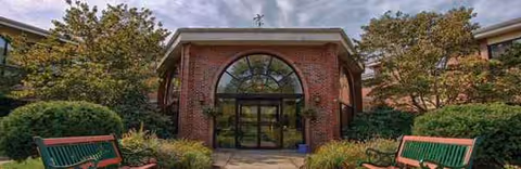 Brick senior living facility entrance with arched glass doors, surrounded by trees and benches.